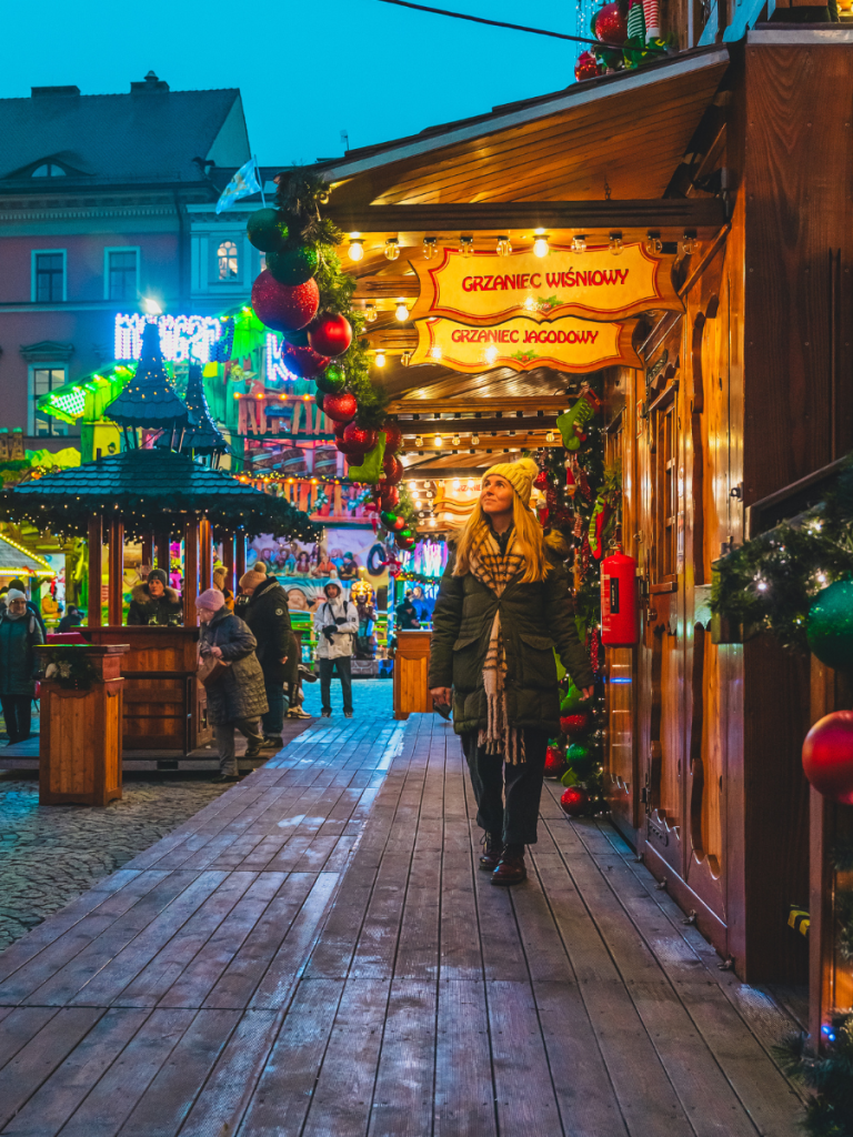 A woman walks past vibrant Christmas stalls adorned with lights and Polish signage in Kraków, surrounded by glowing garlands and marketgoers — soaking in the festive energy of European Christmas markets.