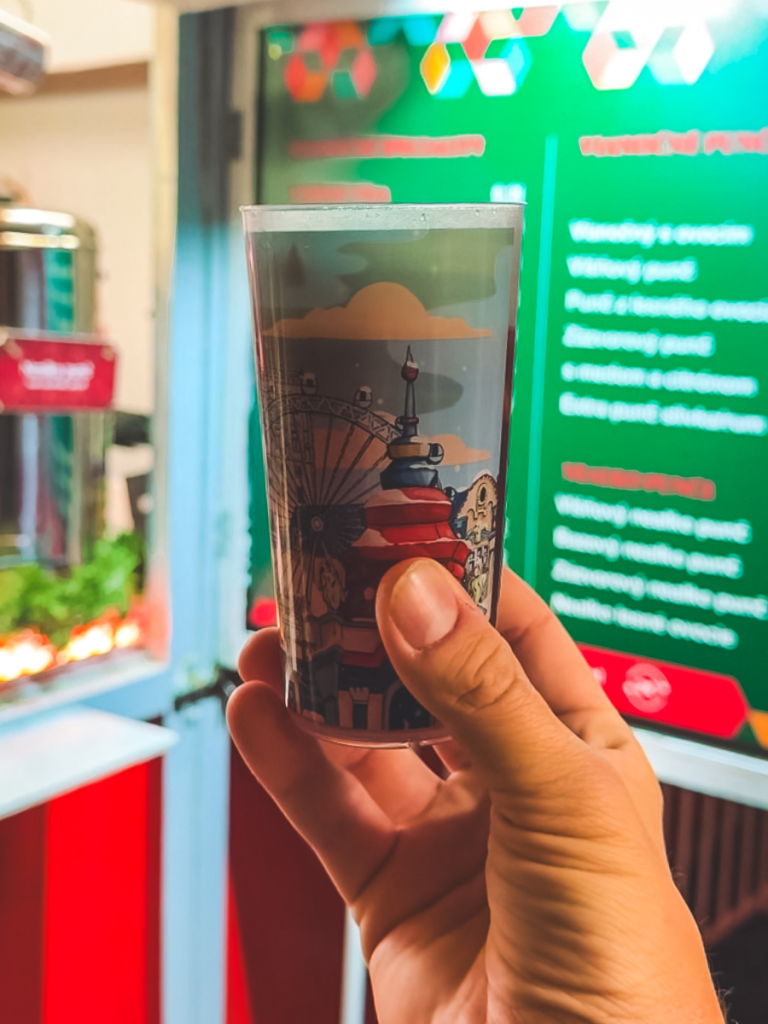 A hand holds a decorated reusable cup filled with hot punch at the Christmas markets in Bratislava, with the festive drink menu visible in the background.