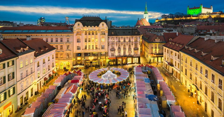 Aerial view of Bratislava’s Christmas market in the main square, with striped red-and-white stalls forming a festive ring of lights below historic buildings and Bratislava Castle glowing in the background — a scenic highlight of European Christmas markets.
