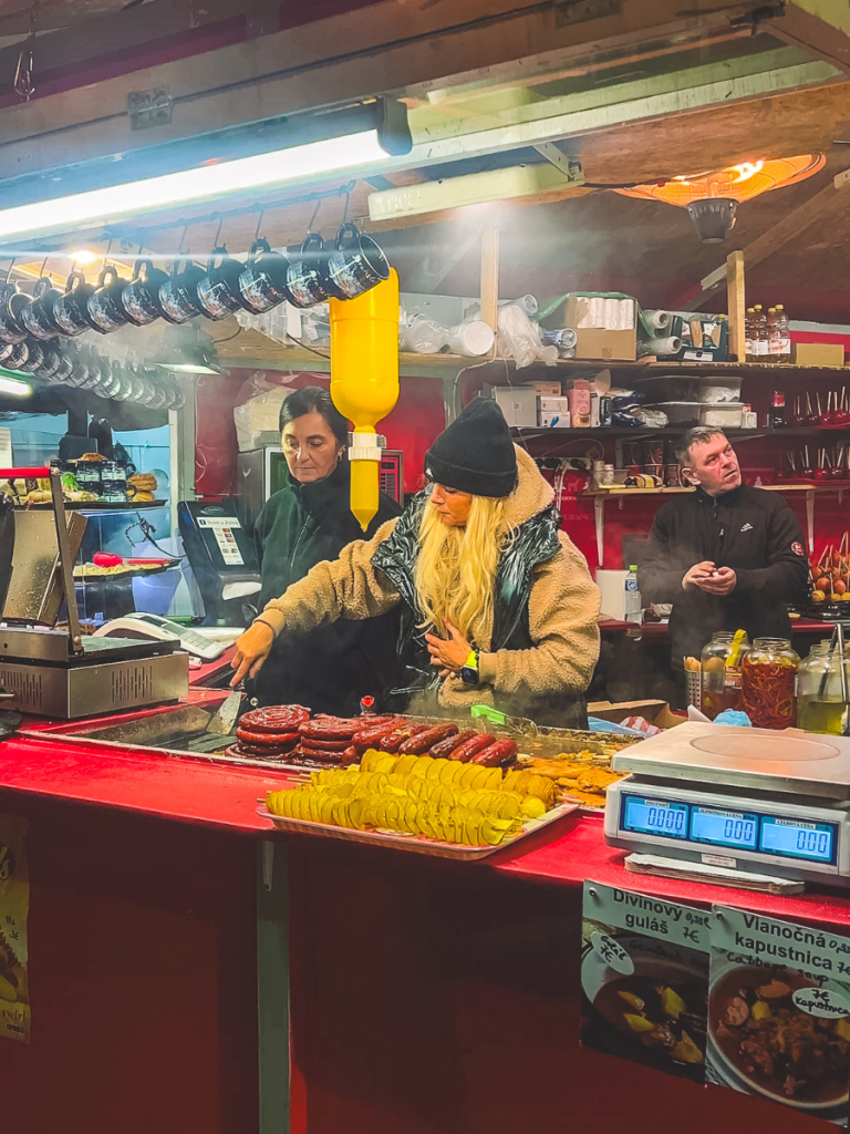 Vendors prepare grilled sausages and spiral potatoes at the Christmas markets in Bratislava, with steam rising and festive mugs hanging above the stall.