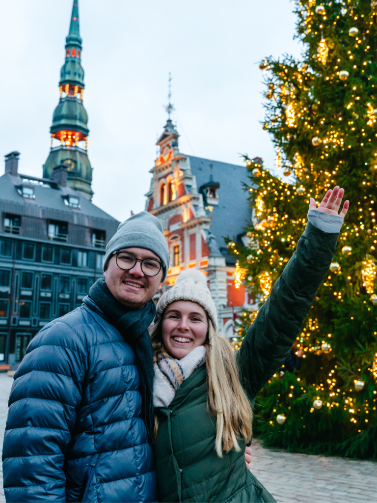 A smiling couple poses beside a glowing Christmas tree in a historic square with festive lights and Gothic buildings — enjoying winter charm in the heart of a European Christmas market.