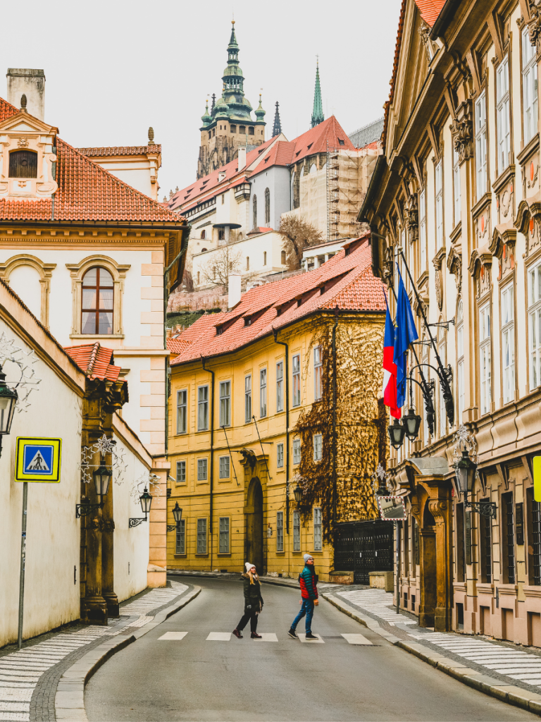 two people crossing the street in prague with prague castle in the background