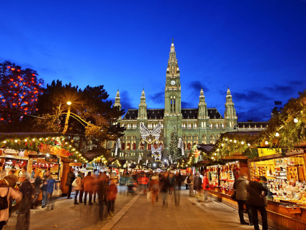 Crowds gather at a brightly lit Christmas market in front of Vienna’s grand Rathaus, with wooden stalls offering warm drinks and treats under twinkling garlands — an iconic evening at one of the most famous European Christmas markets.