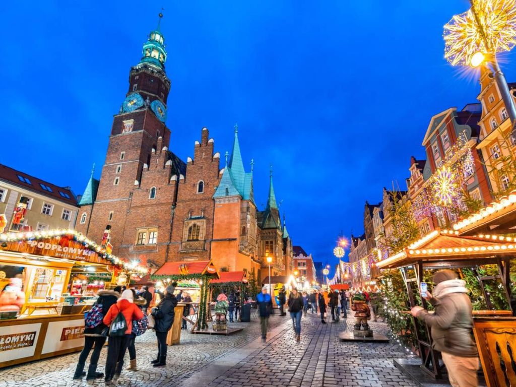 Shoppers explore Wrocław’s festive Christmas market at dusk, with glowing wooden stalls, twinkling lights, and the historic Town Hall towering in the background — a colorful celebration of European Christmas markets in Poland.