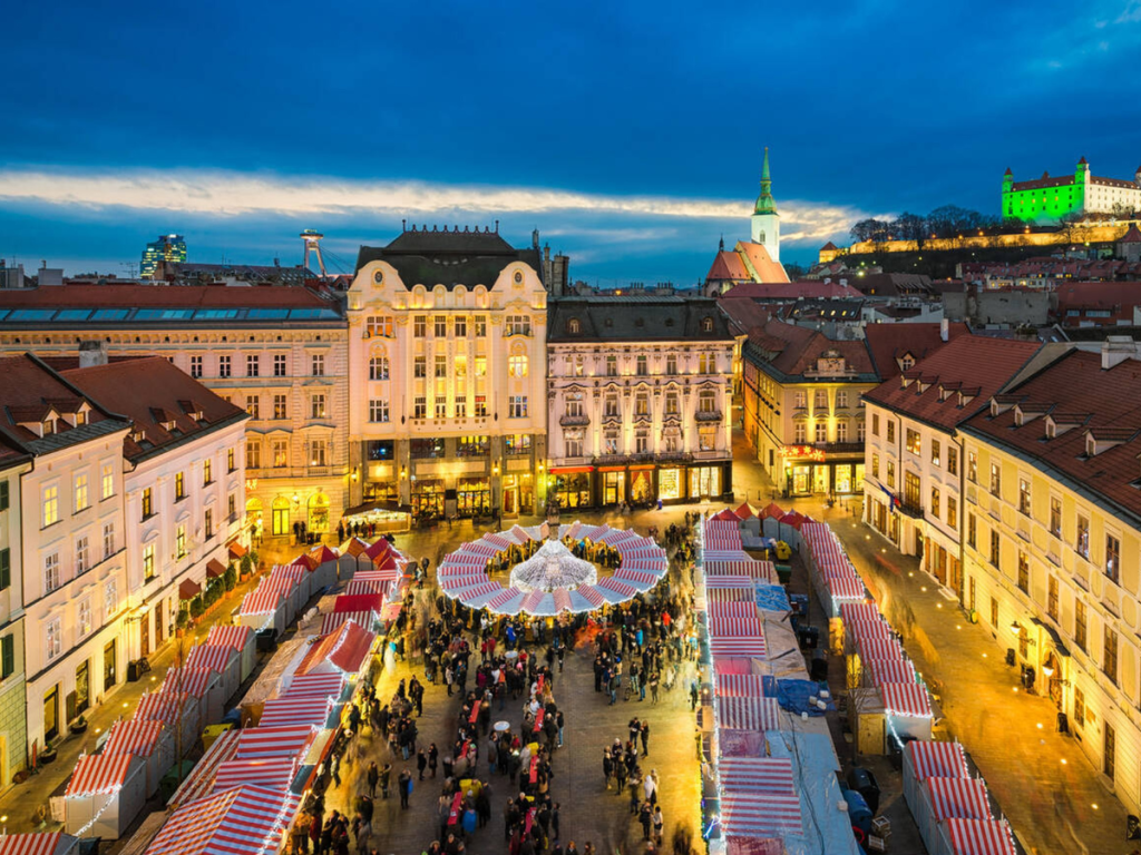 Aerial view of Bratislava’s Christmas market in the main square, with striped red-and-white stalls forming a festive ring of lights below historic buildings and Bratislava Castle glowing in the background — a scenic highlight of European Christmas markets.