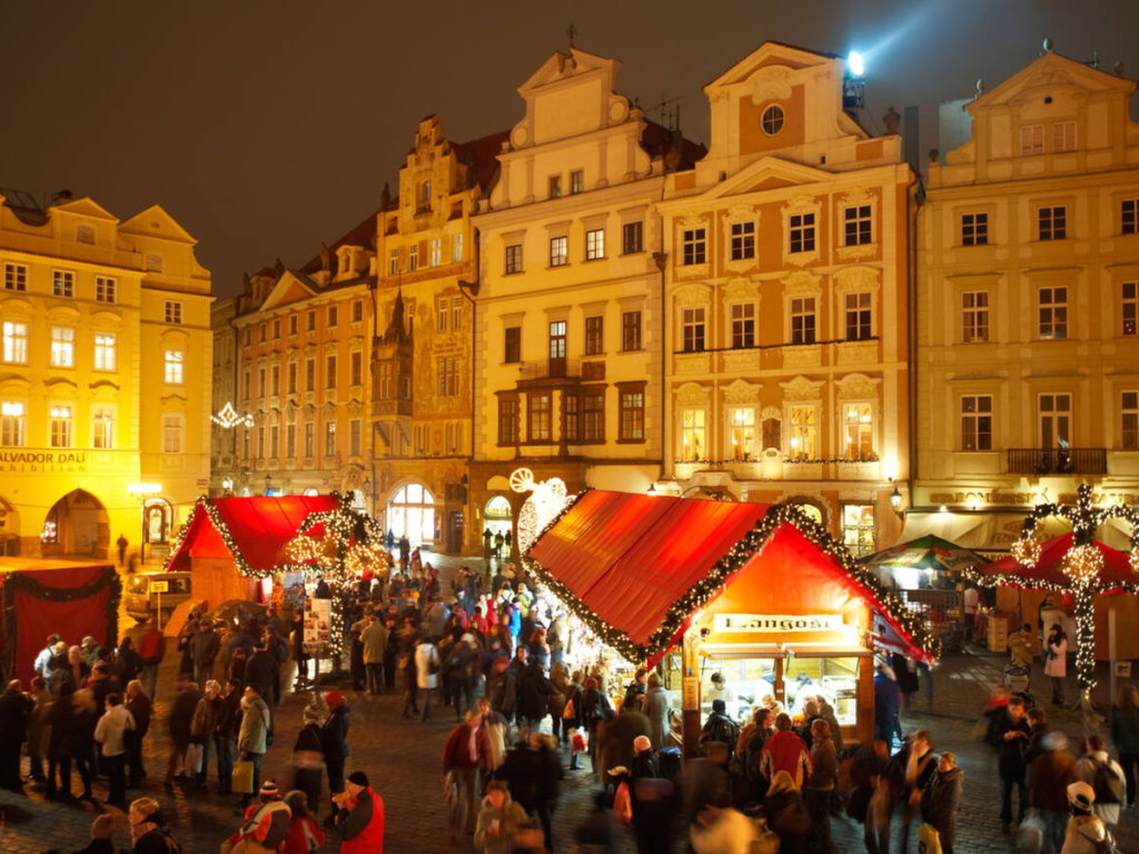 Crowds fill Prague’s Old Town Square at night, exploring red-roofed Christmas market stalls framed by historic pastel buildings and glowing holiday lights — a timeless scene from one of the most beloved European Christmas markets.