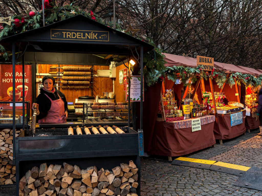 A woman prepares freshly grilled Trdelník pastries at a festive Christmas market stall, surrounded by stacks of firewood, garlands, and booths selling honey wine and sweets — a cozy and flavorful experience at European Christmas markets in the Czech Republic.