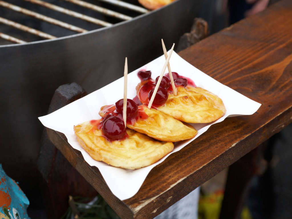 A serving of grilled oscypek cheese topped with sweet berry sauce is presented on a paper tray with toothpicks, resting beside a festive market grill — a traditional and savory snack found at European Christmas markets in Poland.