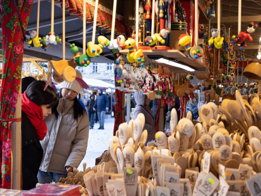 Shoppers browse a colorful Christmas market stall filled with handcrafted wooden spoons, ornaments, and toys, with hanging decorations and traditional embroidered fabric adding festive flair — a joyful scene from European Christmas markets celebrating local artisan goods.