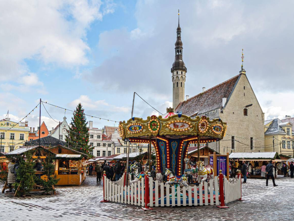 A colorful carousel stands in the center of Tallinn’s snowy Town Hall Square, surrounded by Christmas market stalls, pine trees, and a towering medieval church spire — a magical holiday scene from one of the most picturesque European Christmas markets.