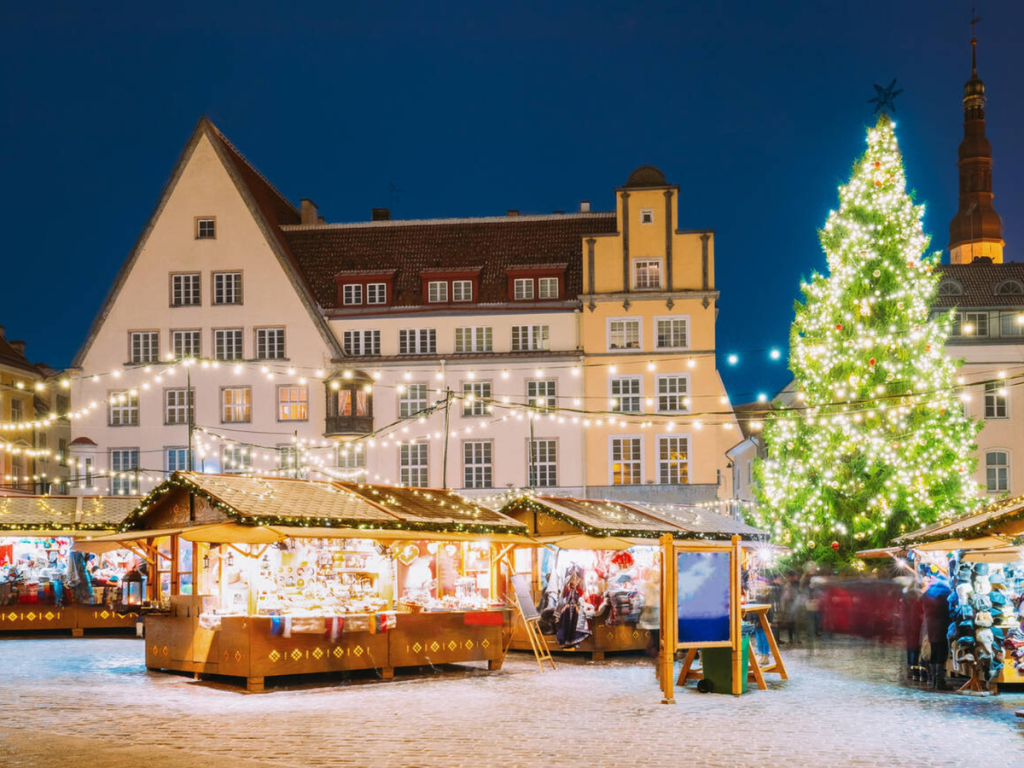 Wooden stalls twinkle under string lights in Tallinn’s Old Town Square as a giant Christmas tree sparkles beside historic buildings — a cozy evening scene at one of the most underrated European Christmas markets.