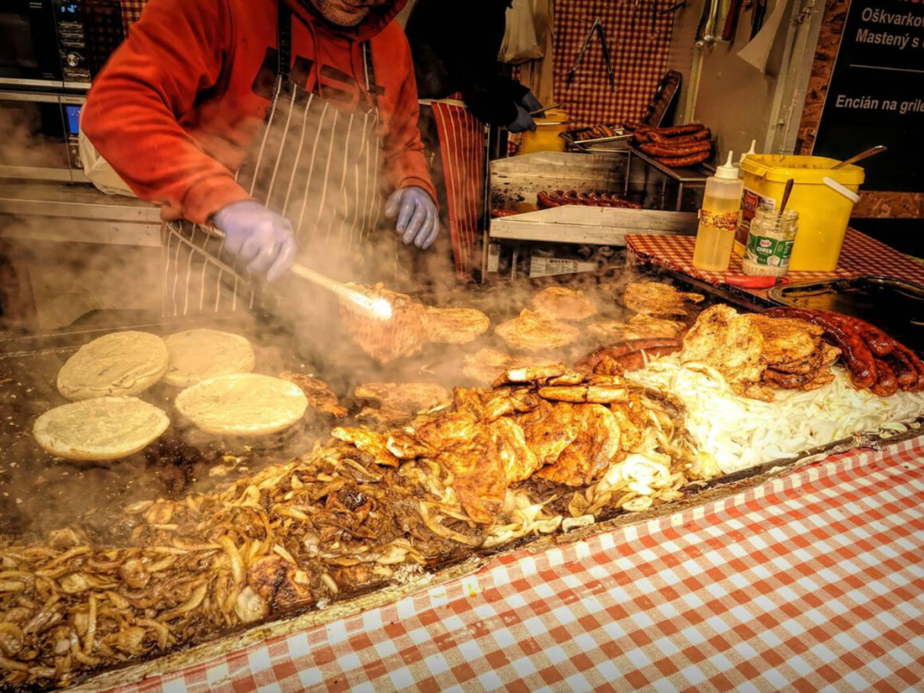 A vendor grilling traditional Slovak street food like sausages, onions, pork, and flatbreads at the Christmas markets in Bratislava, with steam rising over the hot grill.
