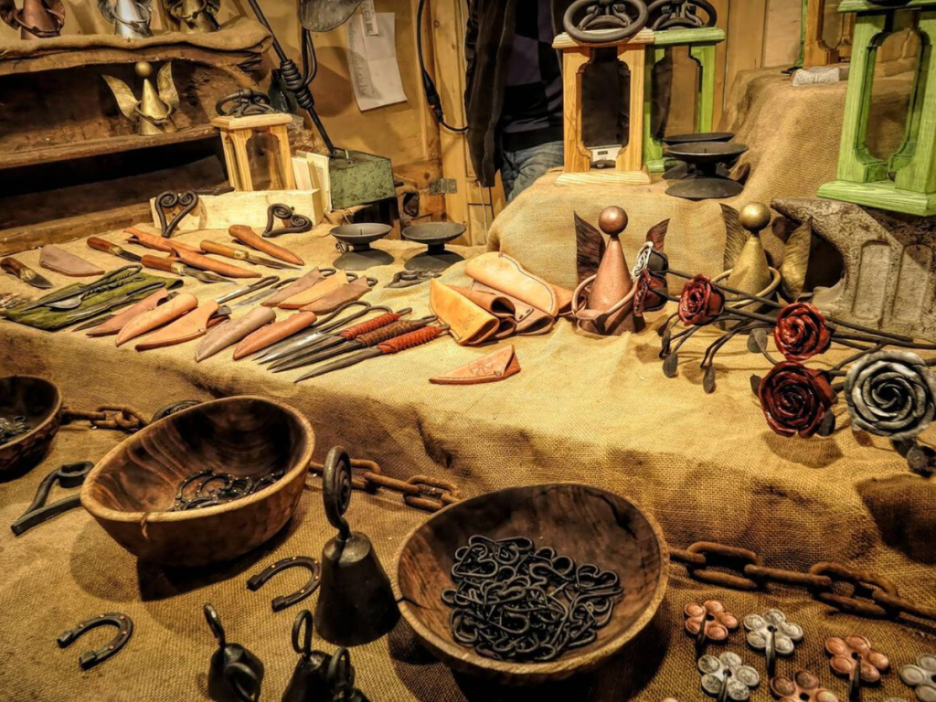 A market stall at the Christmas markets in Bratislava displaying handcrafted metalworks, leather sheaths, forged roses, and decorative gifts on burlap cloth.