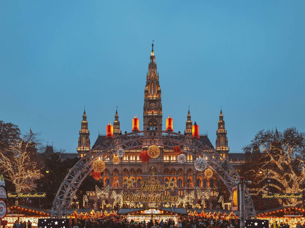 The illuminated entrance to the Vienna Christmas market at Rathausplatz, framed by festive lights and a Gothic-style city hall in the background at twilight.