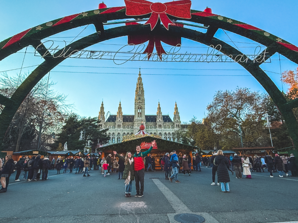 A couple poses under the festive archway of the Wiener Christkindlmarkt with Vienna’s Rathaus in the distance, surrounded by market stalls and holiday crowds on a sunny winter day.