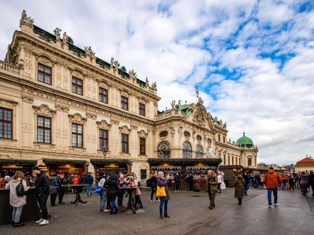 Visitors browse festive stalls and sip mulled wine in front of the ornate façade of Belvedere Palace during the daytime Christmas market in Vienna.