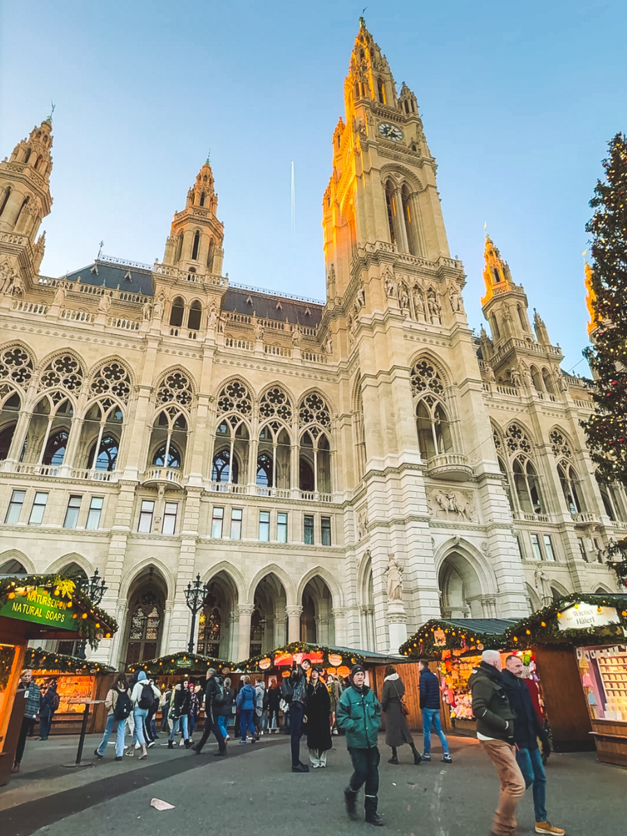 Visitors walking beneath Vienna’s towering Rathaus as golden sunlight illuminates the gothic architecture and festive Christmas market stalls line the square below.