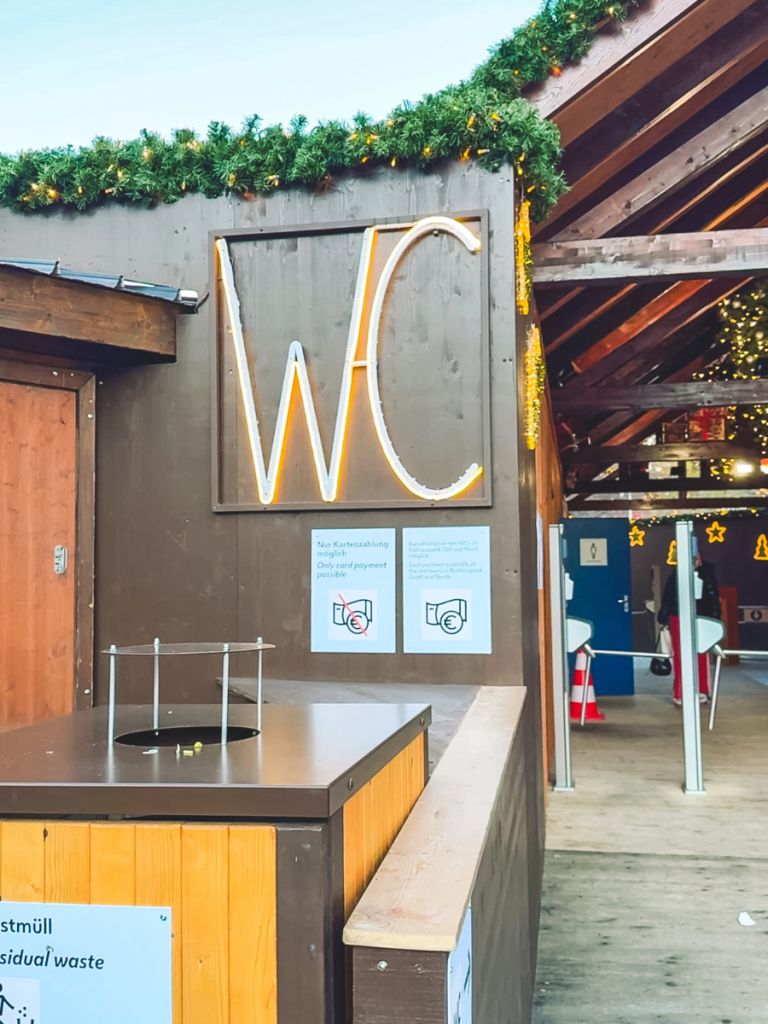 Neon WC sign above the entrance to public restrooms at the Vienna Christmas market, decorated with festive greenery and payment signage for card-only access.