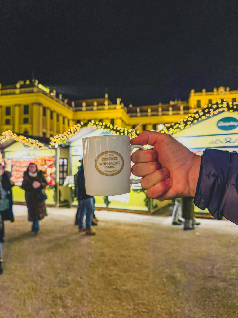 A hand holding a white mug labeled “Weihnachtsmarkt Schönbrunn” in front of festive stalls and twinkling lights at Vienna’s Schönbrunn Palace Christmas Market at night.