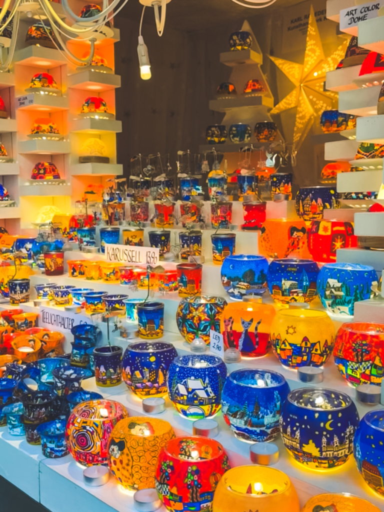 Brightly colored hand-painted glass candle holders displayed at a Vienna Christmas market stall, glowing with tea lights and showcasing festive winter village scenes.
