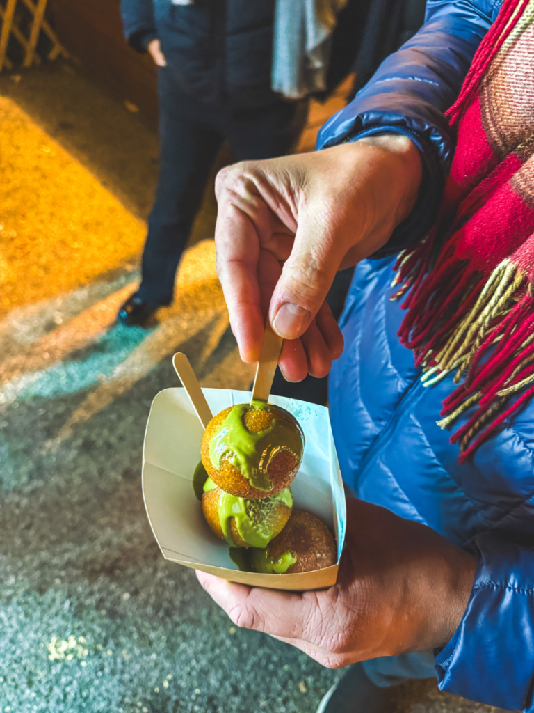 Close-up of a hand holding a skewer of warm donut holes topped with green pistacio glaze, served fresh at a Vienna Christmas market food stall.
