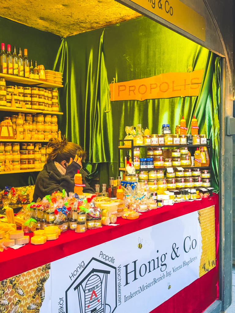 A Christmas market stall in Vienna selling honey, propolis, beeswax candles, and natural skincare products, with jars and gift sets neatly arranged on red tablecloths under a green velvet backdrop.