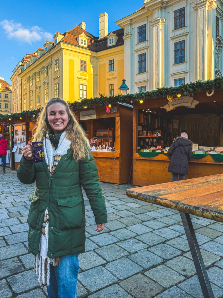 A woman in a green coat smiling while holding a festive mug at the Am Hof Christmas Market in Vienna, surrounded by wooden stalls, bar tables, and pastel-hued historic buildings.