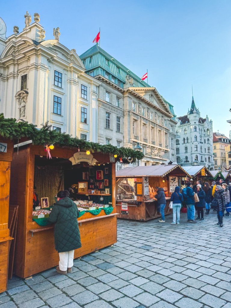 A visitor browses handmade art at a wooden stall at the Am Hof Christmas Market in Vienna, with Austrian flags waving above elegant Baroque and Renaissance-style buildings in the background.