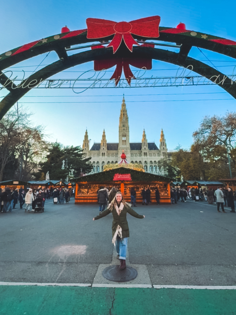 A smiling visitor poses beneath the festive bow-topped archway of the Wiener Christkindlmarkt in Vienna, with the glowing Rathaus and Christmas stalls in the background at twilight.