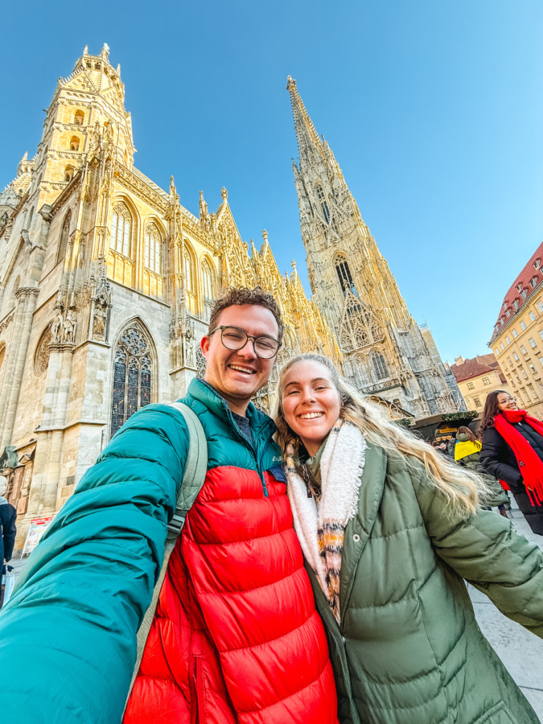 A happy couple takes a selfie in front of the towering gothic spires and golden-lit stone façade of St. Stephen’s Cathedral in Vienna on a clear blue winter day.
