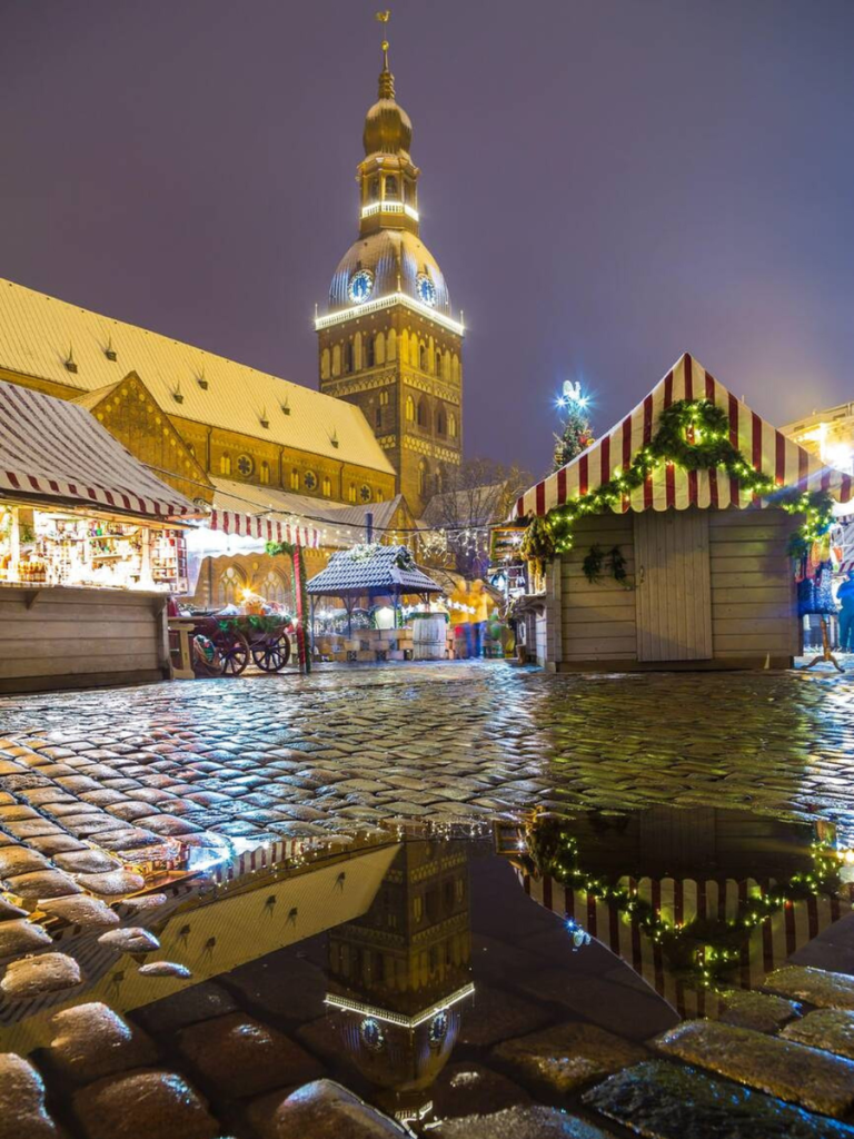 A glowing church tower reflects in a puddle on the cobblestone streets of Riga’s Christmas market, surrounded by striped stalls, twinkling lights, and light snowfall — a dreamy winter evening at one of Europe’s most atmospheric Christmas markets.