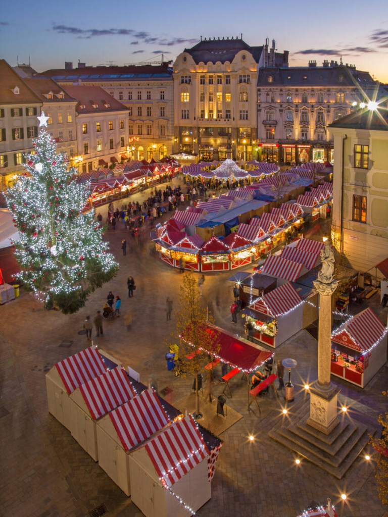 A festive aerial view of the Christmas markets in Bratislava’s main square, with red-and-white striped stalls, a giant lit Christmas tree, and historic buildings at dusk.