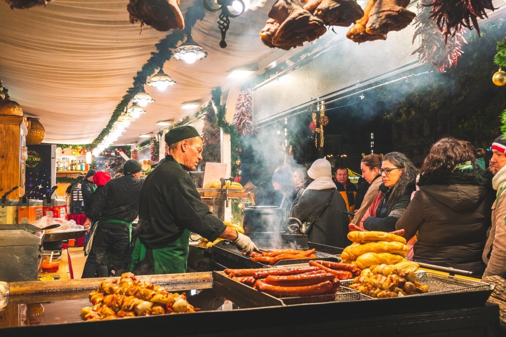 A chef grills sausages and skewers at a lively Christmas market food stall, with steam rising as visitors line up under festive garlands and hanging meats — a deliciously authentic moment at a European Christmas market.