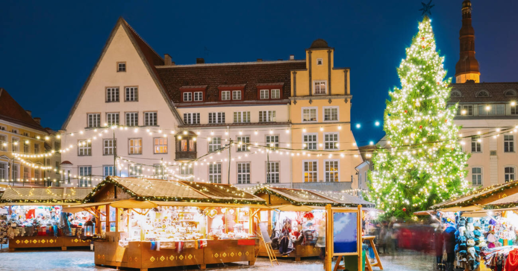 Wooden stalls twinkle under string lights in Tallinn’s Old Town Square as a giant Christmas tree sparkles beside historic buildings — a cozy evening scene at one of the most underrated European Christmas markets.