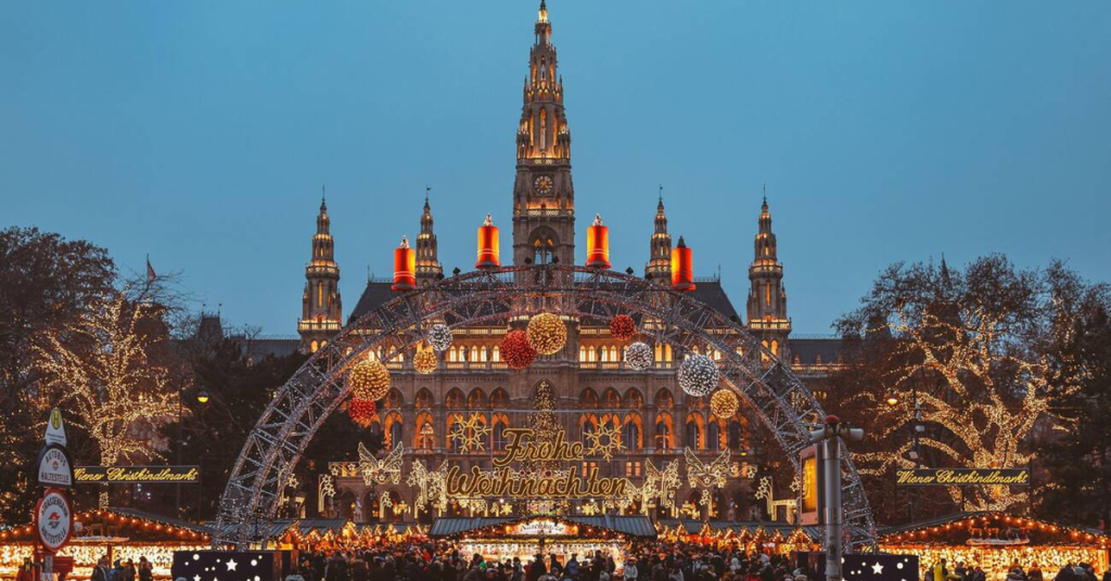 The festive archway and glowing "Frohe Weihnachten" sign at Vienna’s Rathausplatz Christmas Market, with the illuminated Gothic Rathaus building and decorated trees creating a magical holiday scene at twilight.