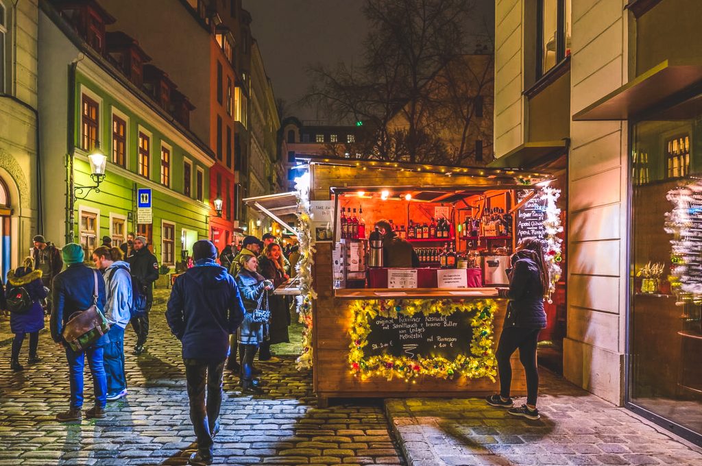 People gather around a warmly lit wooden Glühwein stall tucked into a charming cobblestone street in Vienna, surrounded by colorful buildings and twinkling holiday lights.