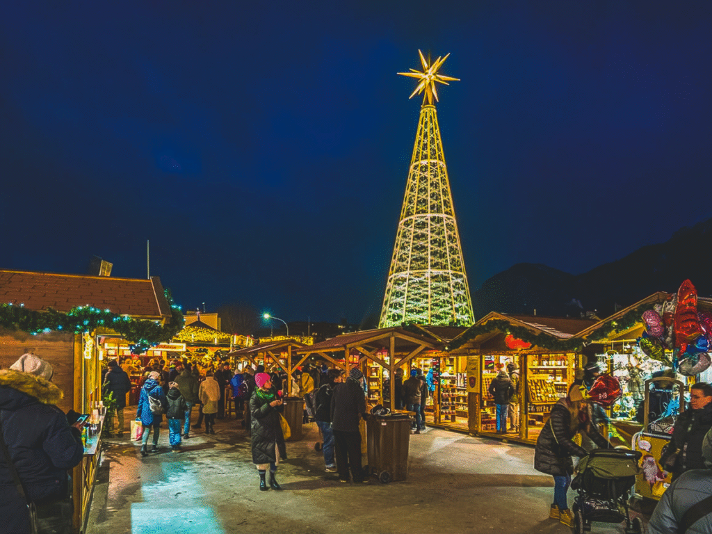 Visitors walking through the festive Christmas market in Innsbruck, Austria, with wooden stalls, twinkling lights, and a towering illuminated tree at night