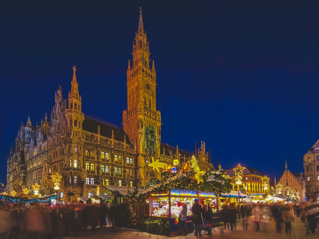 Crowds browsing Christmas stalls at Munich’s Christkindlmarkt in Marienplatz, with festive lights and the illuminated New Town Hall tower in the background