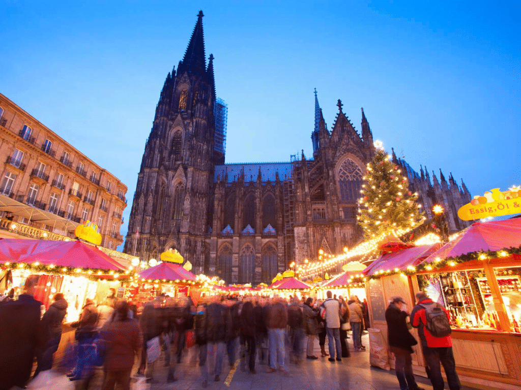 Crowds enjoying the Christmas market in front of the Cologne Cathedral in Germany, with red-roofed stalls, festive lights, and a large Christmas tree