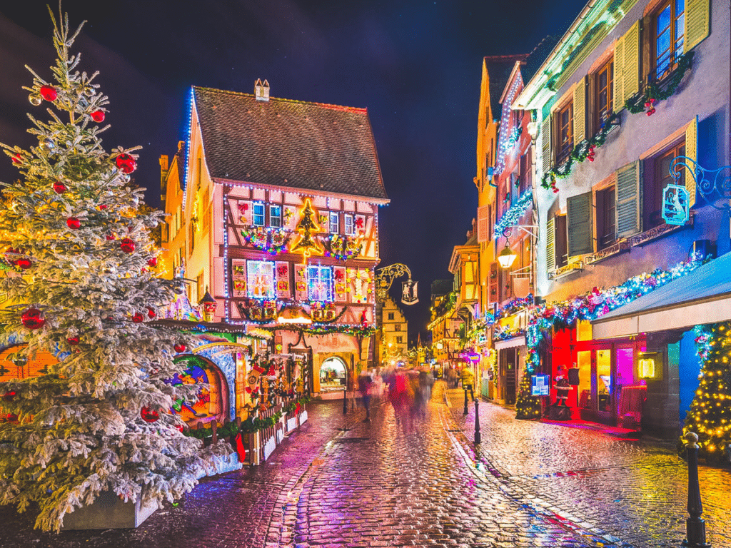 Charming Christmas scene in Colmar, France, with colorful half-timbered houses, festive lights, and a snow-dusted Christmas tree along cobblestone streets