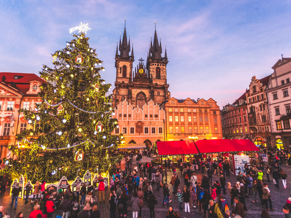 Crowds gathering around a giant illuminated Christmas tree at Prague’s Old Town Square with historic buildings and the Church of Our Lady before Týn in the background, featured as a must-visit stop on a European Christmas market itinerary.