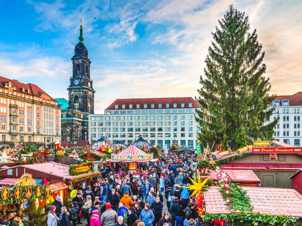 Crowds exploring the colorful wooden stalls, giant Christmas tree, and festive decorations at the Dresden Striezelmarkt, with the historic church tower in the background, featured as a must-see stop on a European Christmas market itinerary.