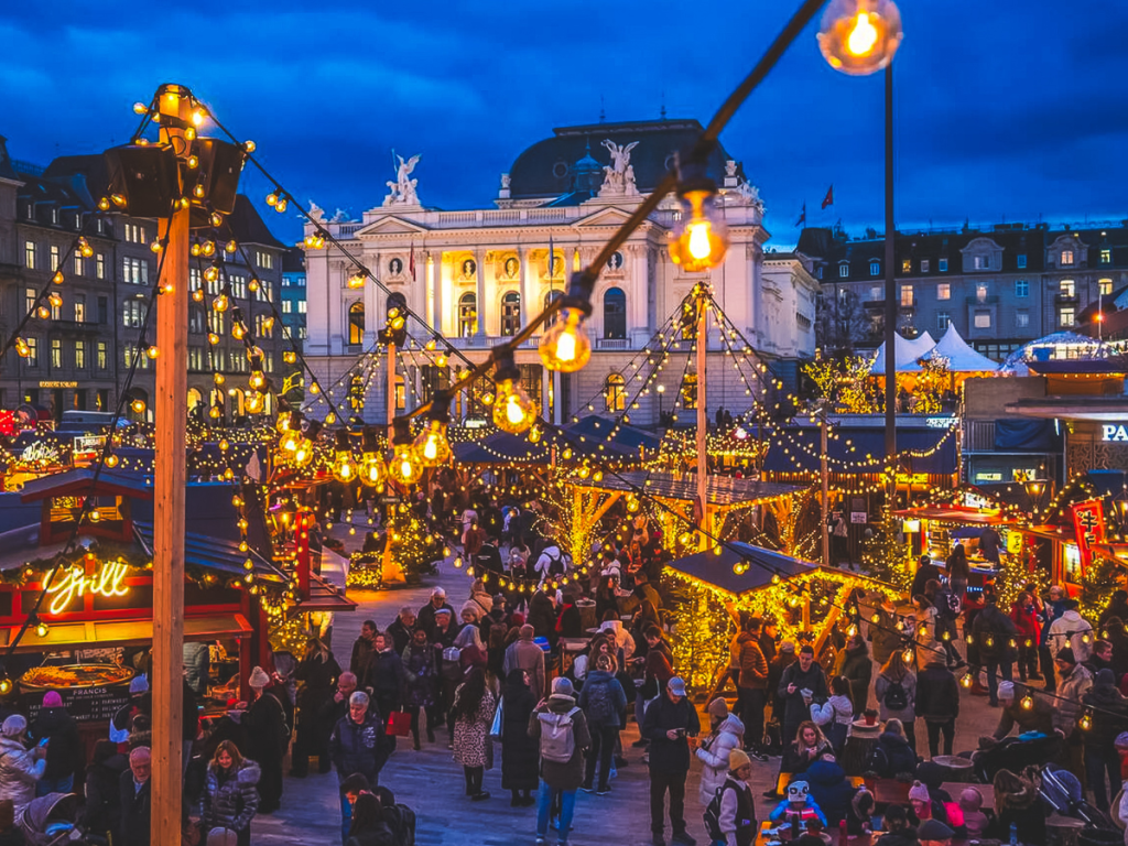 Crowds enjoying the festive Christmas market in front of the illuminated Zurich Opera House, surrounded by glowing string lights, decorated stalls, and holiday trees, highlighted as a magical stop on a European Christmas market itinerary.