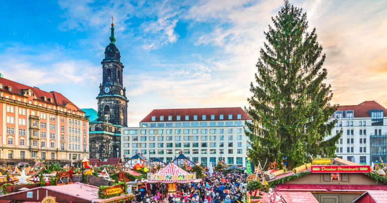 Crowds exploring the colorful wooden stalls, giant Christmas tree, and festive decorations at the Dresden Striezelmarkt, with the historic church tower in the background, featured as a must-see stop on a European Christmas market itinerary.
