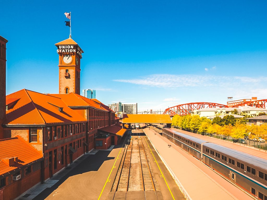 A bright daytime view of Portland Union Station showcasing the historic clock tower, train platforms, and nearby red steel bridge under clear blue skies.