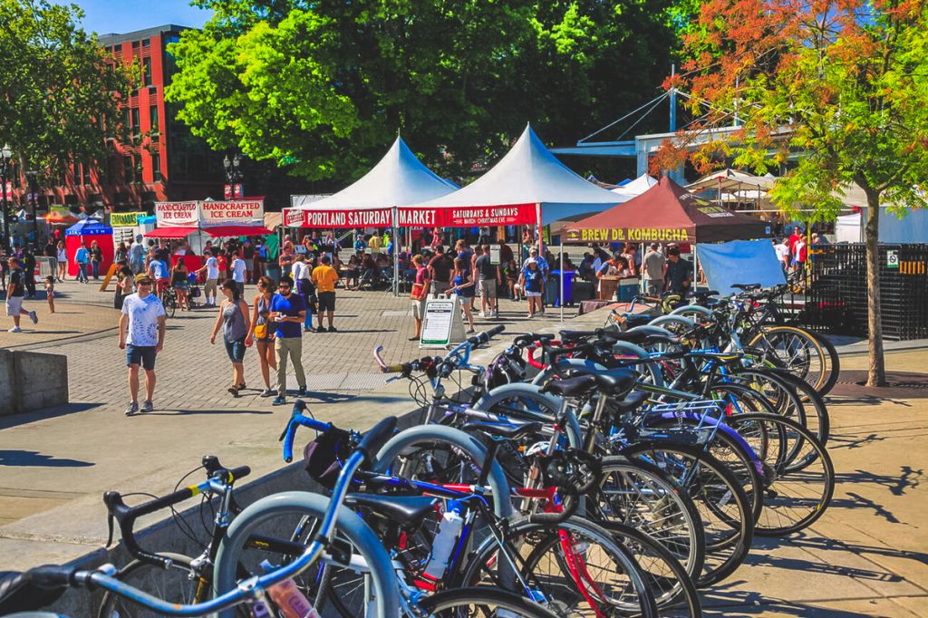 Bikes lined up near the Portland Saturday Market with vendor tents, crowds, and trees along the waterfront