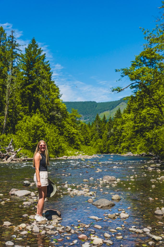 Woman standing on rocks in a shallow river surrounded by lush green trees and forested hills on a sunny day