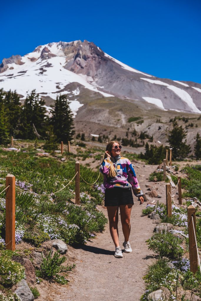 Woman walking along a wildflower-lined trail near Timberline Lodge with Mount Hood rising in the background on a sunny day