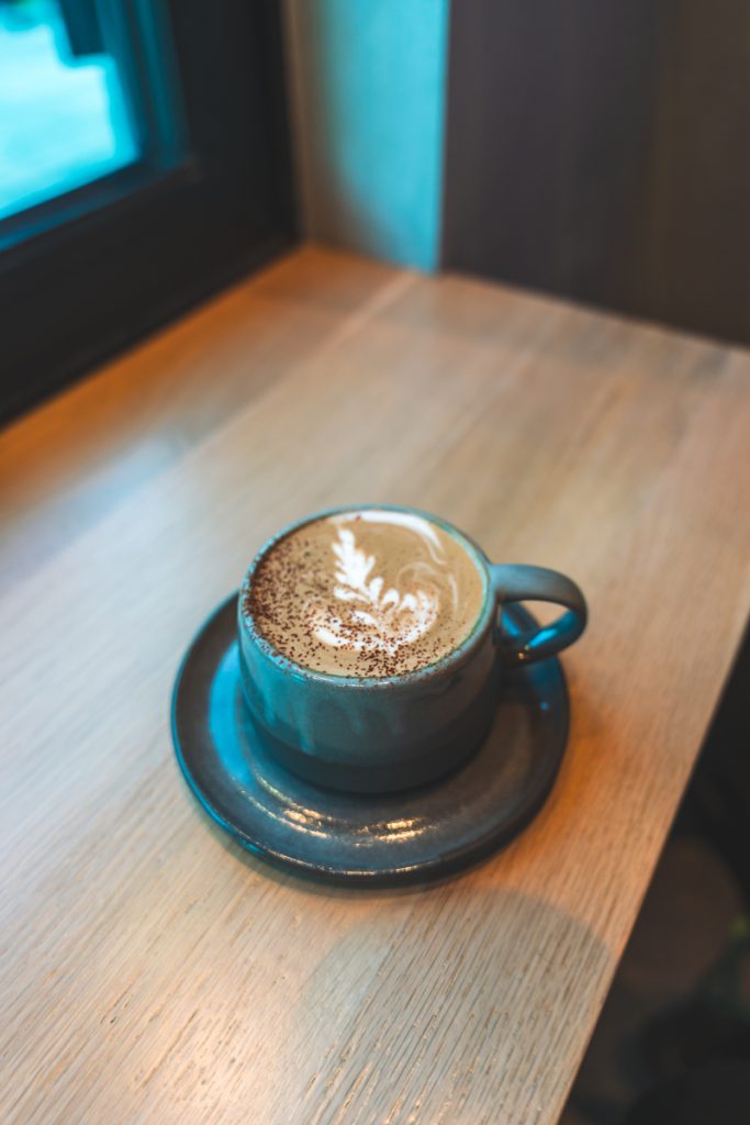 Ceramic cup of latte with leaf latte art sitting on a wooden table near a window