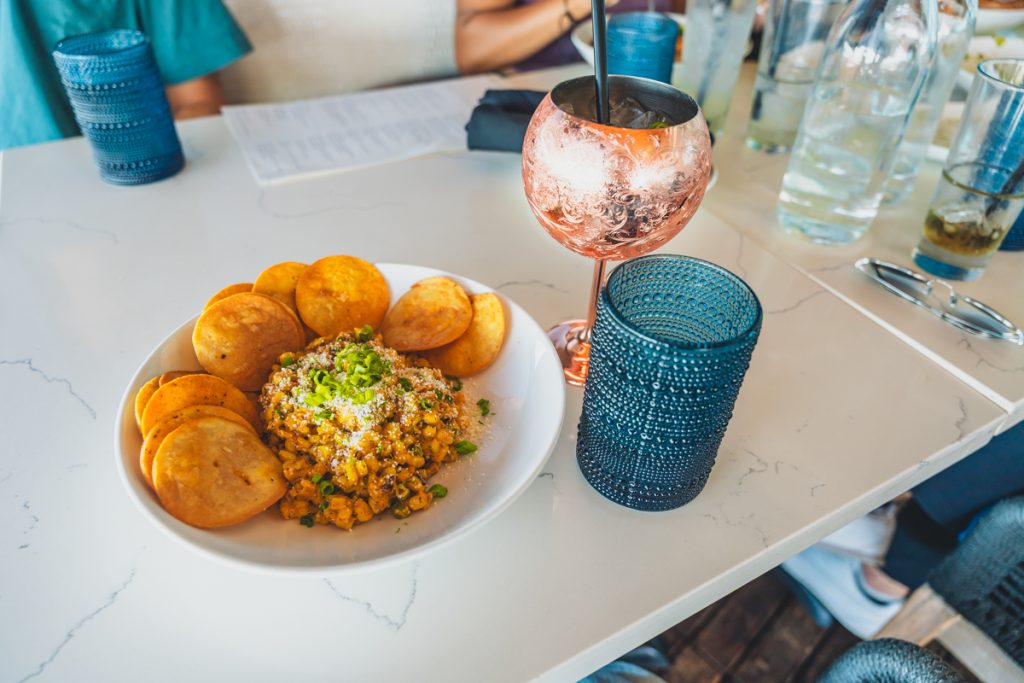 Plate of elotes topped with cheese and herbs served with crispy chips and a cocktail on a restaurant table at Dosalas